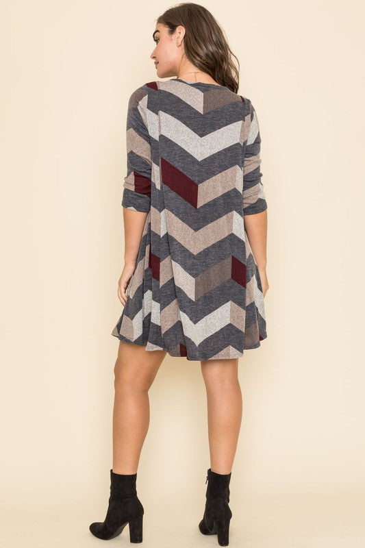 A person stands against a plain background, showcasing the Plus Chevron A-Line Dress in grey, taupe, and teal. The dress features three-quarter sleeves and is crafted from a polyester spandex blend, effortlessly combining comfort and style.