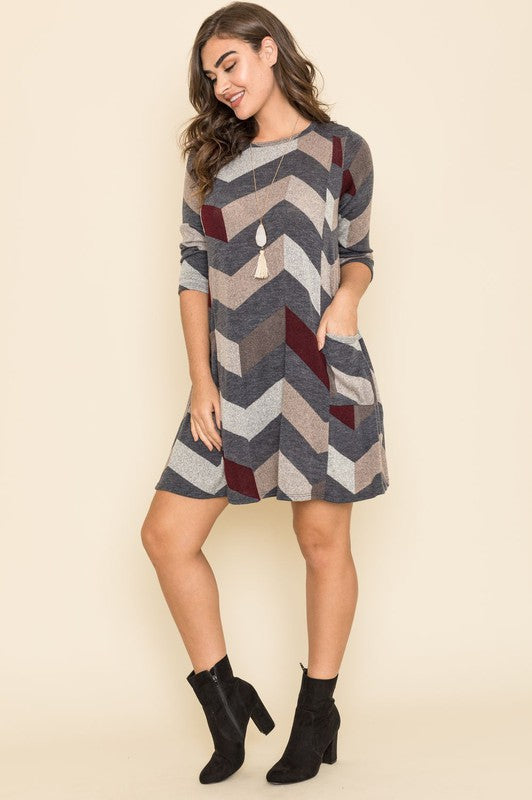 A person stands against a plain background, wearing the Plus Chevron A-Line Dress. The dress, featuring shades of gray, beige, and teal with an elegant chevron pattern, boasts long sleeves and an above-the-knee length. Crafted from fine fabrics and proudly Made in the United States.