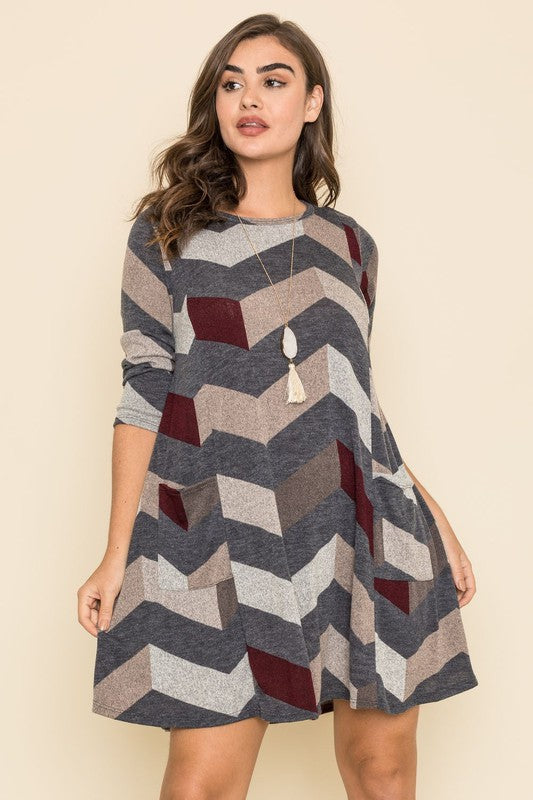 A person stands against a plain background, showcasing the Plus Chevron A-Line Dress in grey, taupe, and teal. The dress features three-quarter sleeves and is crafted from a polyester spandex blend, effortlessly combining comfort and style.