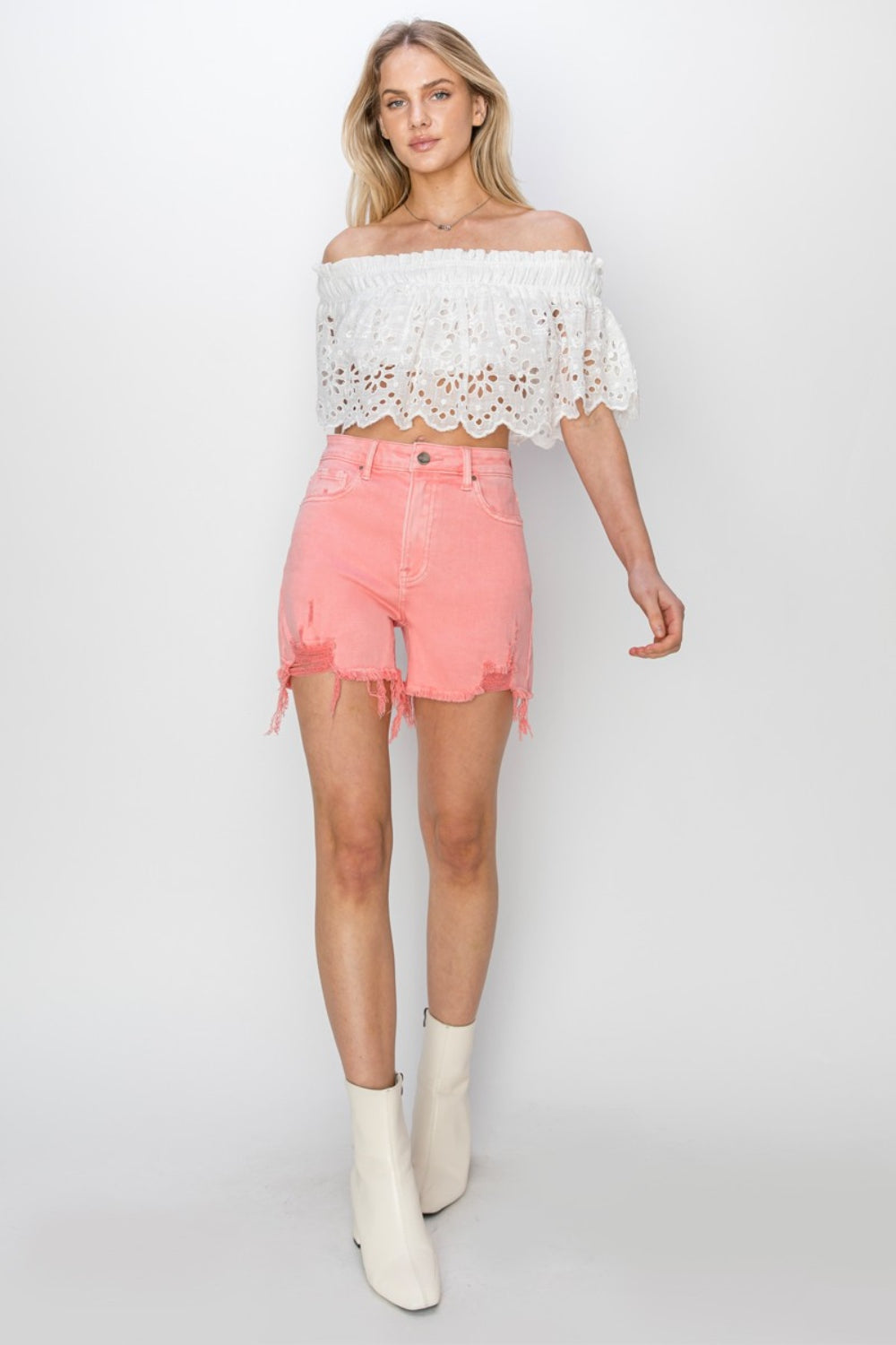 Sporting a stylish look with RISEN High Rise Distressed Denim Shorts in frayed pink and a white lace top, someone stands against a plain backdrop. The distressed details of the denim add an edgy flair to this chic outfit.