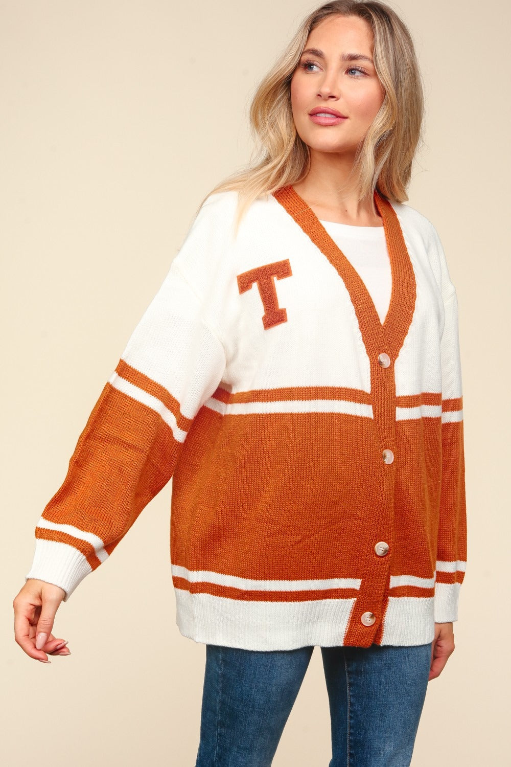 A smiling woman wearing the Haptics Full Size V Neck Button Down Letter Patch Cardigan, an oversized fit in white and orange with a "T" patch, poses with one hand on her hip and the other touching her hair.