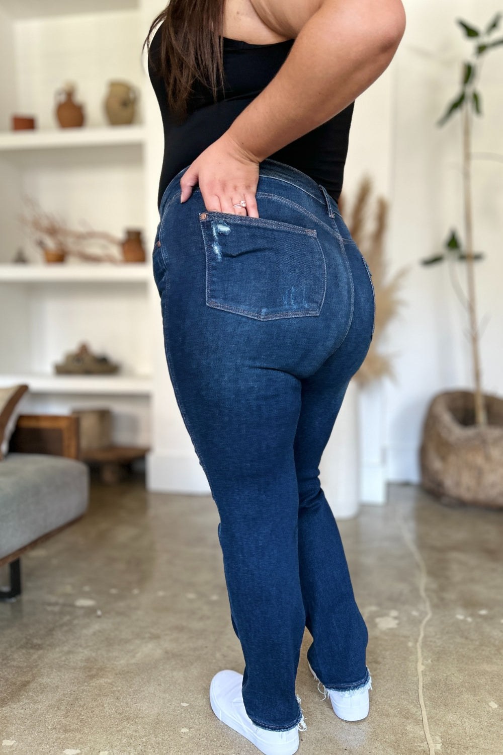 A woman in a black sleeveless top and Judy Blue Full Size High Waist Rigid Magic Heavy Destroy Straight Jeans stands indoors with her hands in her pockets. The background features a plant and shelves adorned with decor items.