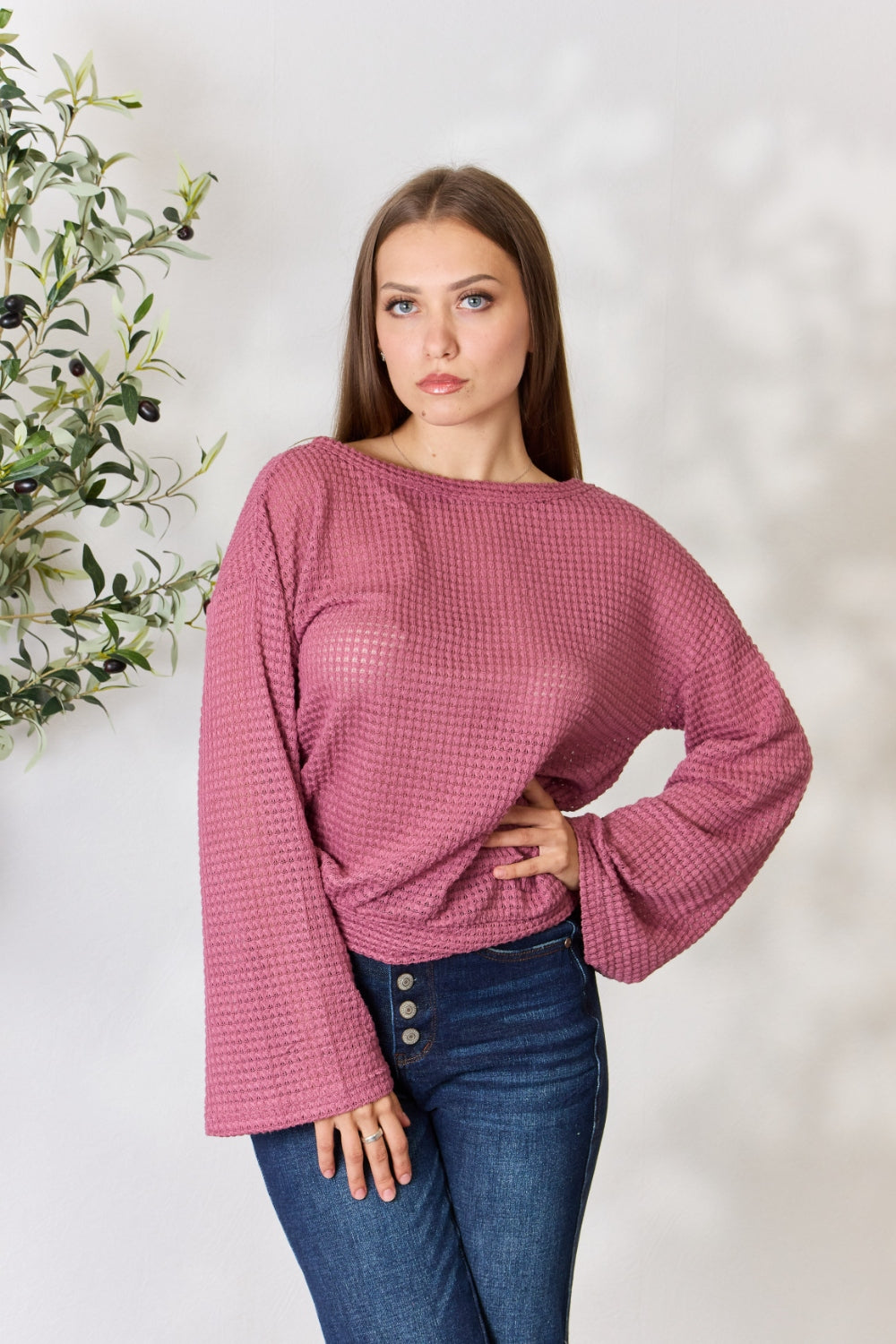 A person wearing a Culture Code Full Size Waffle-Knit Round Neck Long Sleeve Blouse in pink and jeans stands in front of a light background, complemented by a leafy plant on the left.