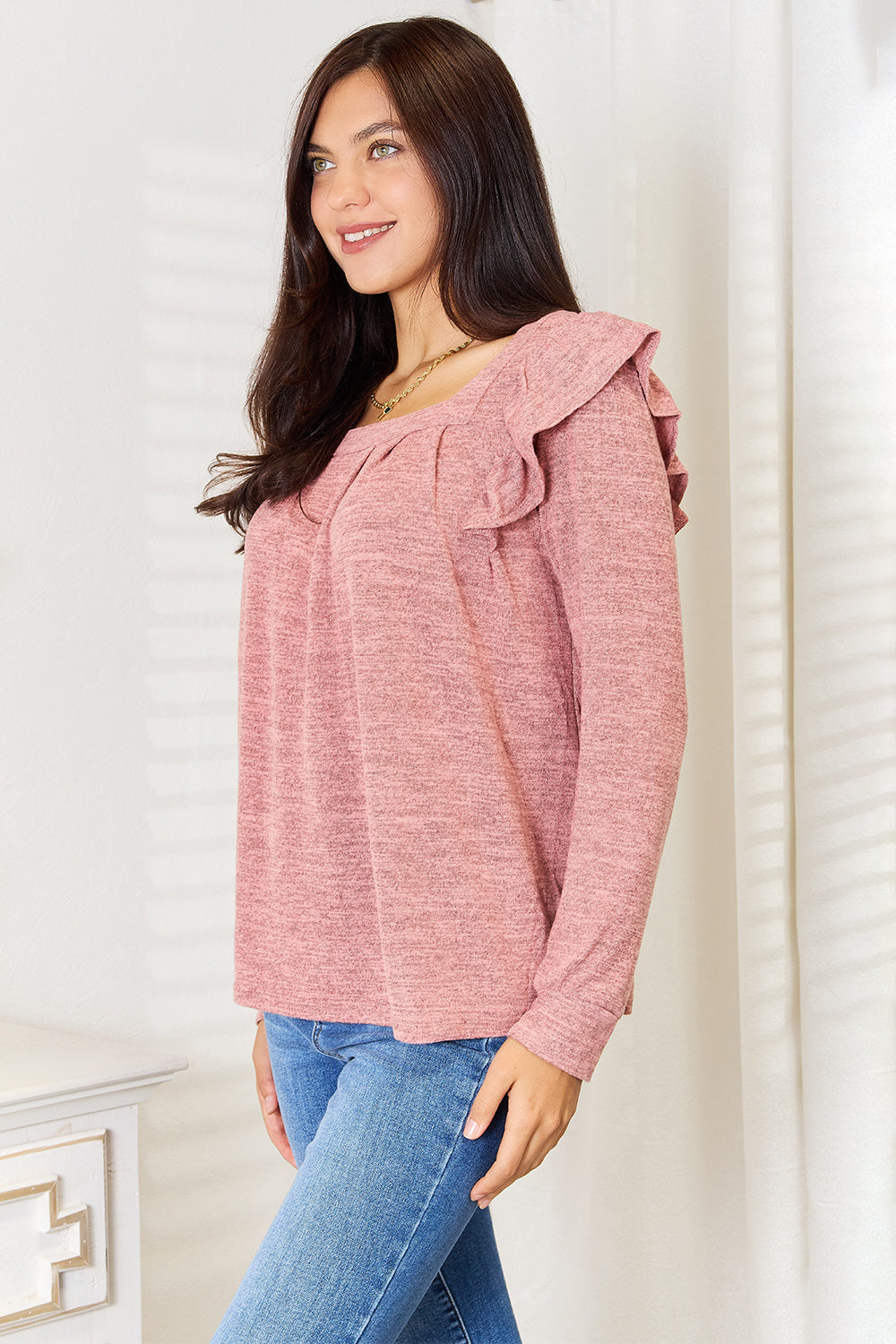 A woman wearing a Double Take Square Neck Ruffle Shoulder Long Sleeve T-Shirt in pink and blue jeans stands with one hand in her pocket, smiling. She has long curly hair and wears a necklace and bracelet. She stands in a room with white decor.