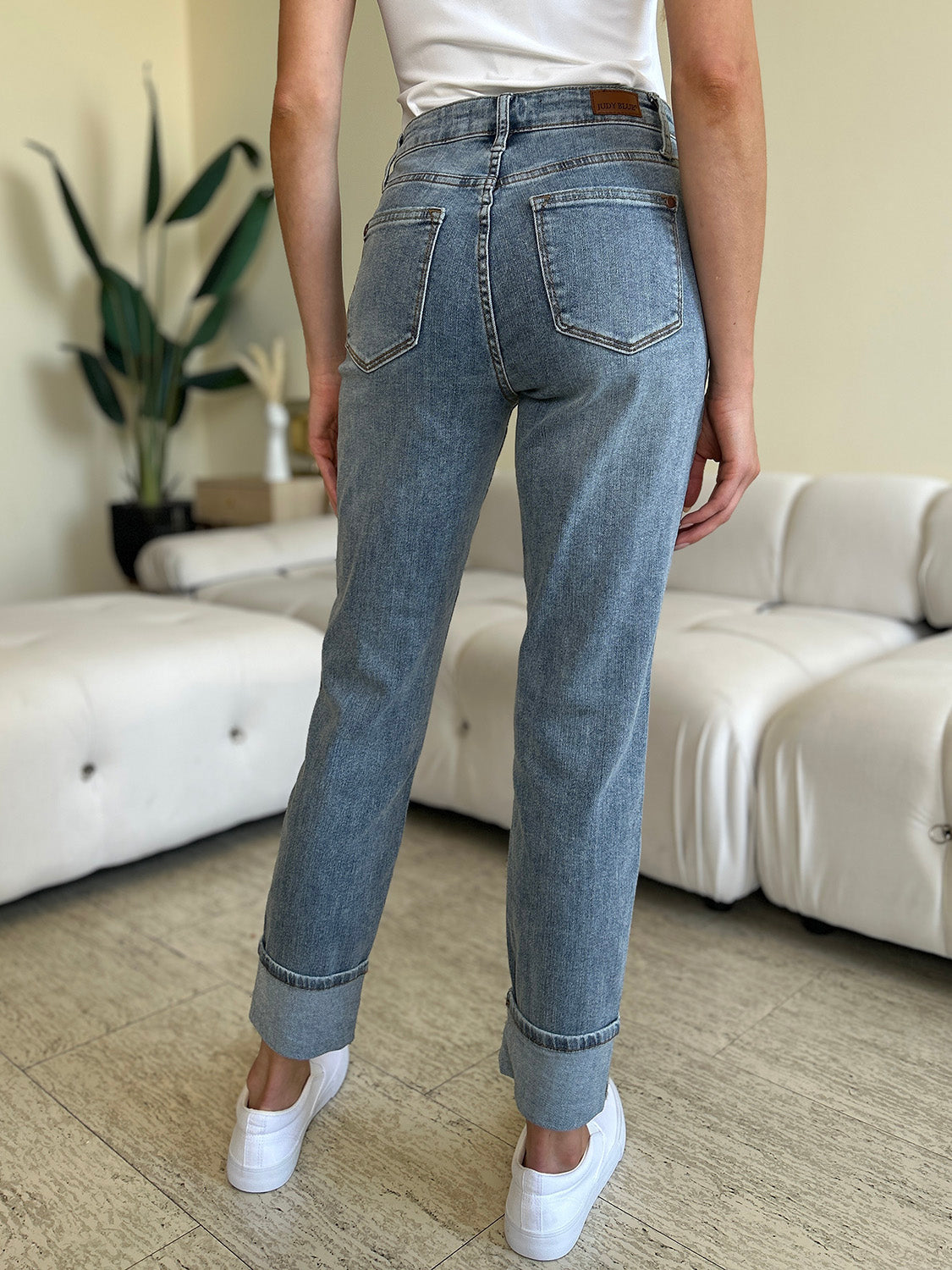 A person wearing Judy Blue Full Size High Waist Cuff Hem Jeans and white slip-on shoes stands in a living room with a white couch and plants in the background.