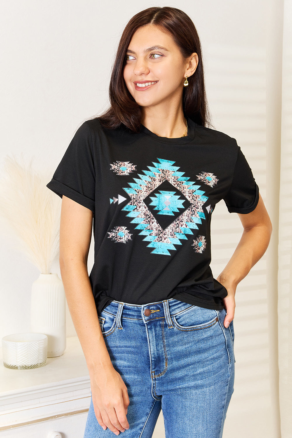 A woman stands indoors wearing the Simply Love Graphic Short Sleeve T-Shirt, a stylish black tee featuring a striking blue and white geometric pattern. Made from comfortable fabric, she has one hand on her hip and is smiling.