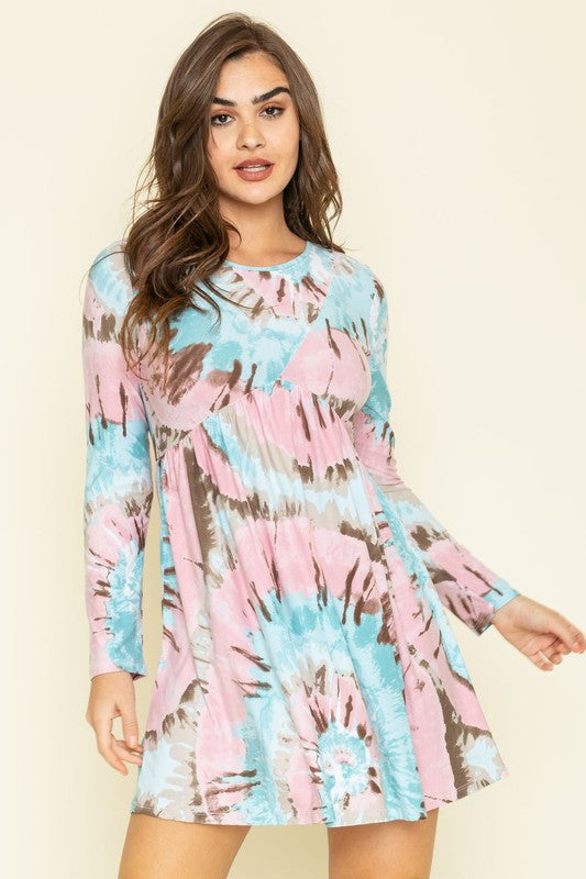 Against a beige background, a woman smiles radiantly as she sports the Cross Swirl Tie Dye Mini Dress with long sleeves. Made in the United States, this outfit embodies a perfect fusion of style and comfort.