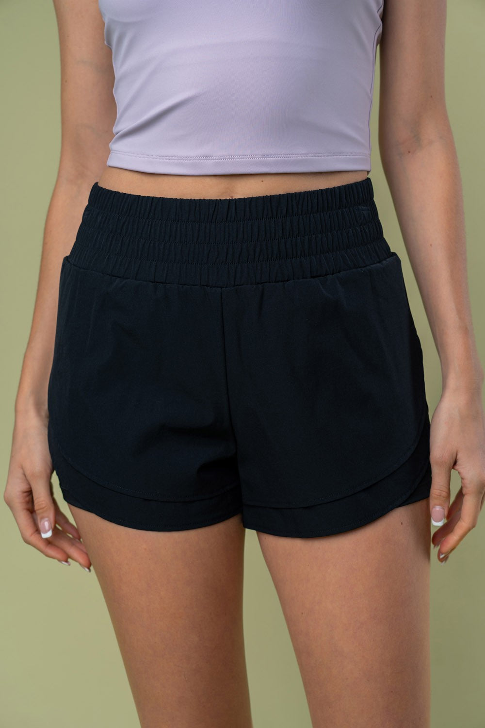 A person wearing a light-colored crop top and White Birch Full Size High Waisted Knit Shorts with an elastic waistband, placing a hand in the side pocket of the shorts against a plain background.