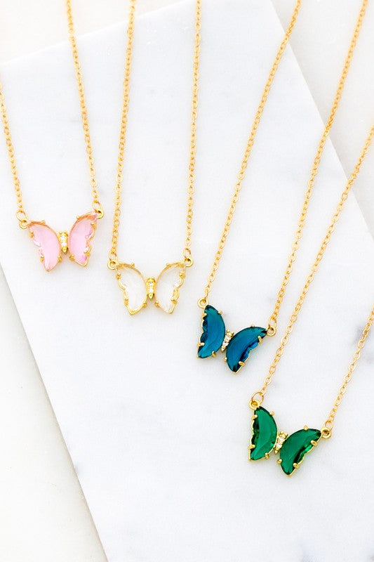 The Gem Stone Butterfly Pendant Necklace, available in an assortment of pastel hues and adorned with exquisite gemstone accents, is featured alongside matching earrings in shades of green, blue, pink, and clear, all elegantly displayed on a white surface.