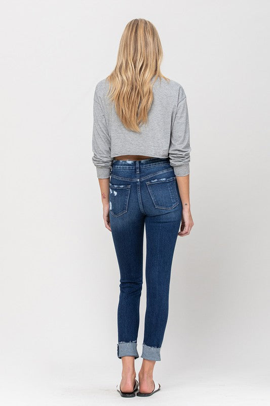 A person wearing a gray cropped sweater paired with High Rise Distressed Clean Cut Crop Skinny blue jeans and white slide sandals stands against a plain background.