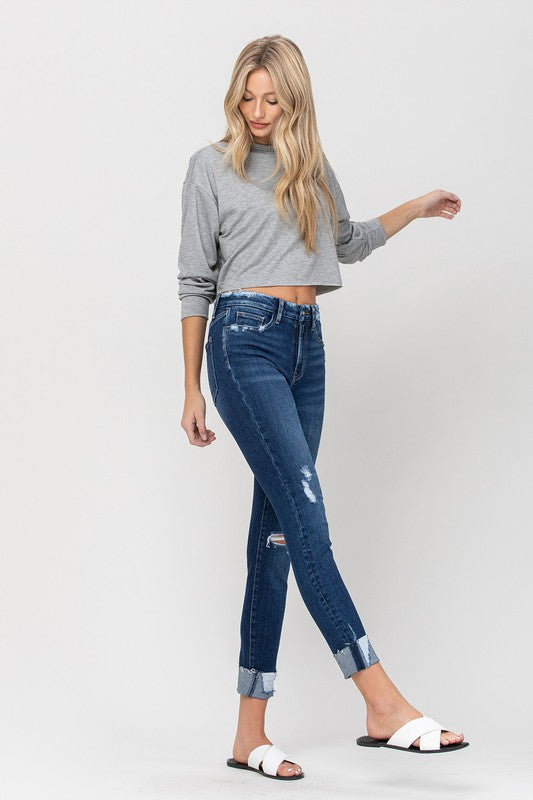 A person wearing a gray cropped sweater paired with High Rise Distressed Clean Cut Crop Skinny blue jeans and white slide sandals stands against a plain background.