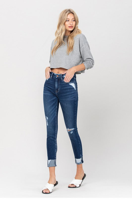 A person wearing a gray cropped sweater paired with High Rise Distressed Clean Cut Crop Skinny blue jeans and white slide sandals stands against a plain background.