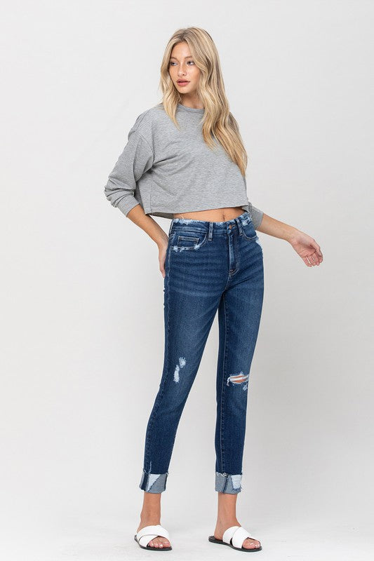 A person wearing a gray cropped sweater paired with High Rise Distressed Clean Cut Crop Skinny blue jeans and white slide sandals stands against a plain background.