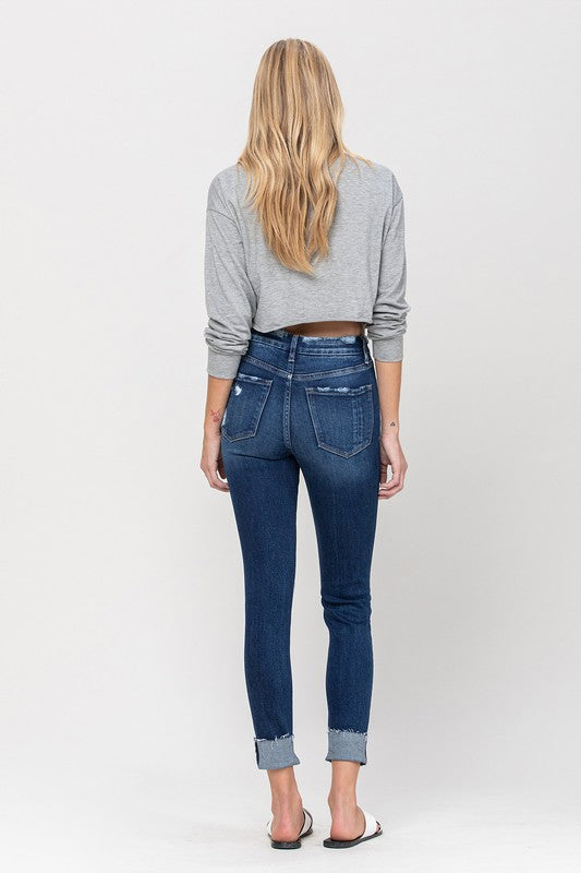 A person wearing a gray cropped sweater paired with High Rise Distressed Clean Cut Crop Skinny blue jeans and white slide sandals stands against a plain background.