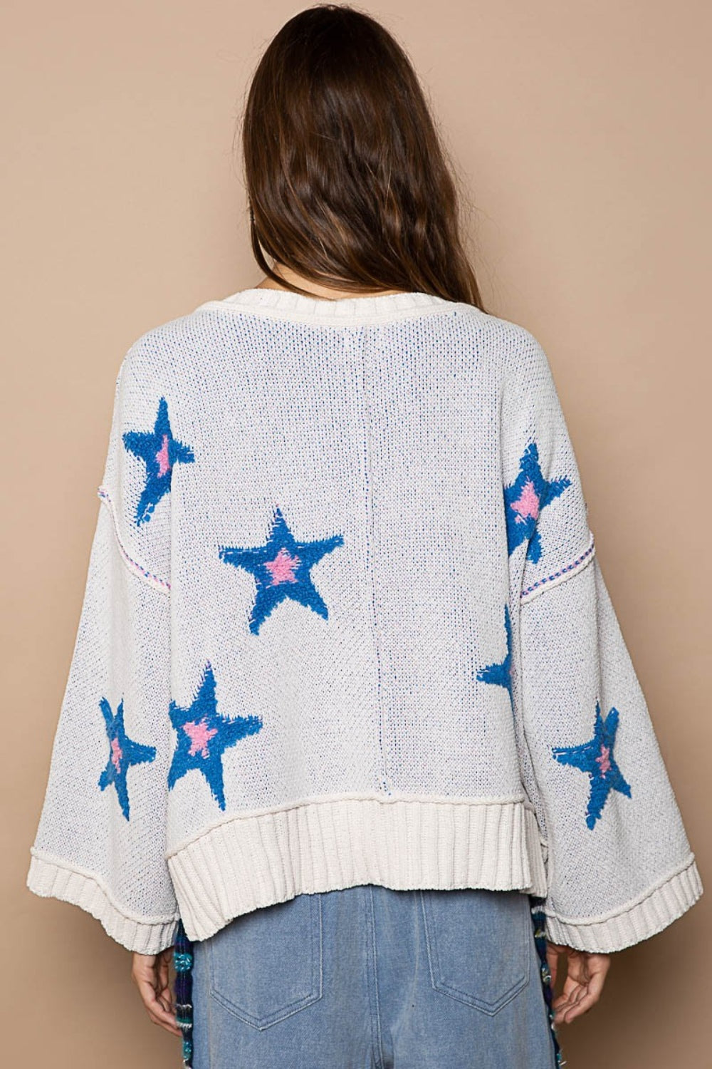 Woman in a POL Long Sleeve Star Patch Sweater with blue color-blocked star berber patches and light blue jeans standing against a beige background.