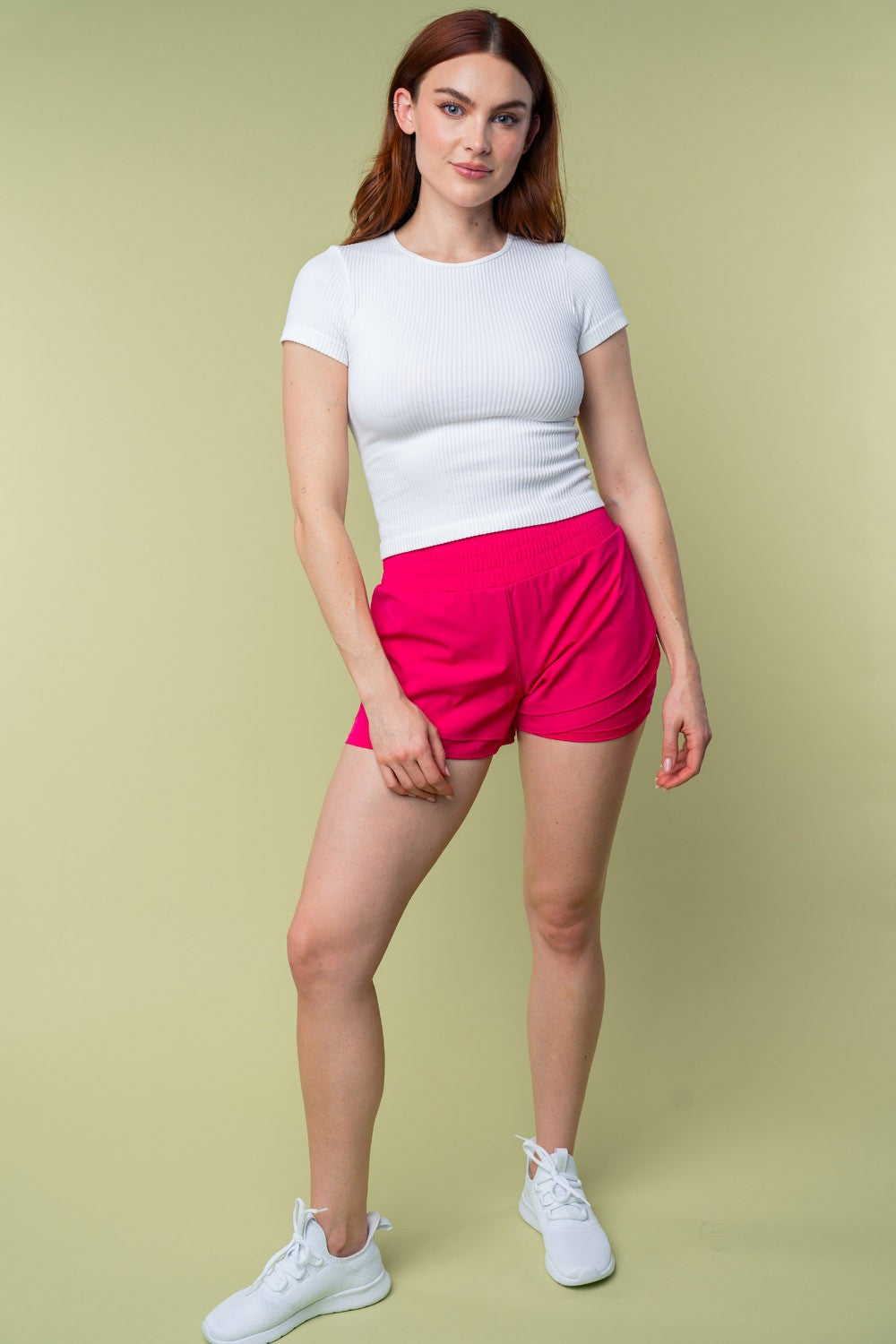 A person wearing White Birch Full Size High Waisted Knit Shorts in bright pink and a white cropped top stands against a light green background.