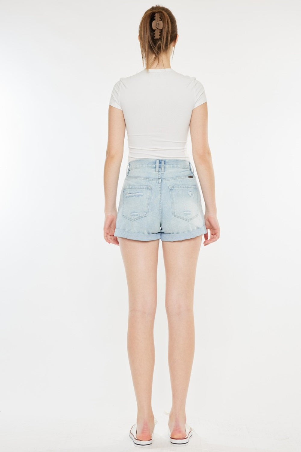 A person stands against a plain background, wearing a white short-sleeve crop top, Kancan High Rise Repaired Mom Denim Shorts in light blue, and white sneakers.