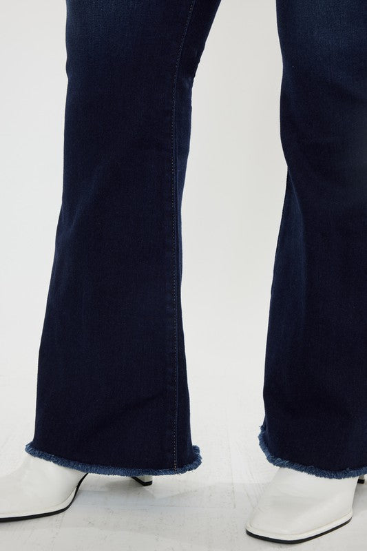 A person stands against a white background, dressed in a black t-shirt and white shoes, showcasing the flattering fit of the Plus High Rise Double WB Fray Hem Flare Jeans in dark blue stretch denim.