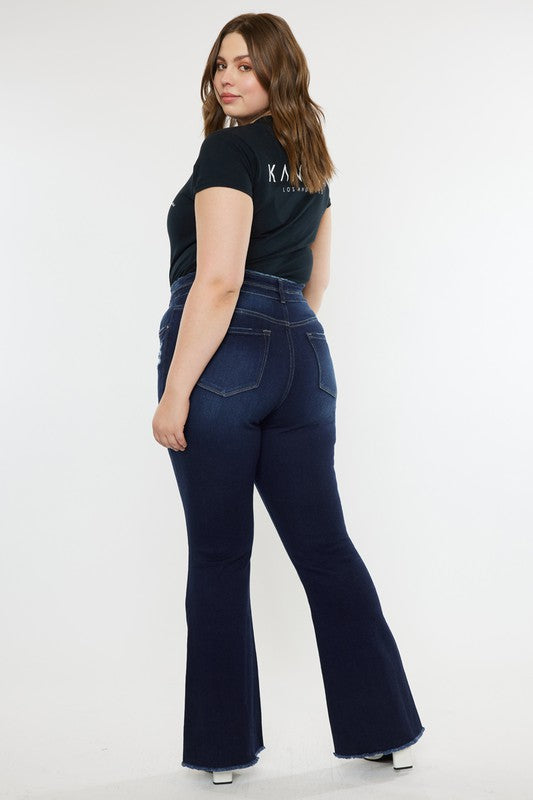 A person stands against a white background, dressed in a black t-shirt and white shoes, showcasing the flattering fit of the Plus High Rise Double WB Fray Hem Flare Jeans in dark blue stretch denim.