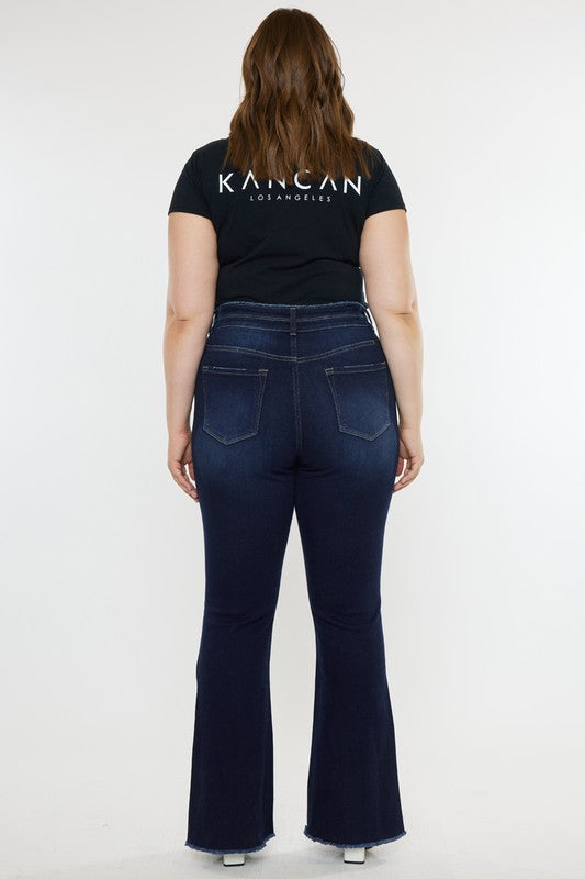 A person stands against a white background, dressed in a black t-shirt and white shoes, showcasing the flattering fit of the Plus High Rise Double WB Fray Hem Flare Jeans in dark blue stretch denim.