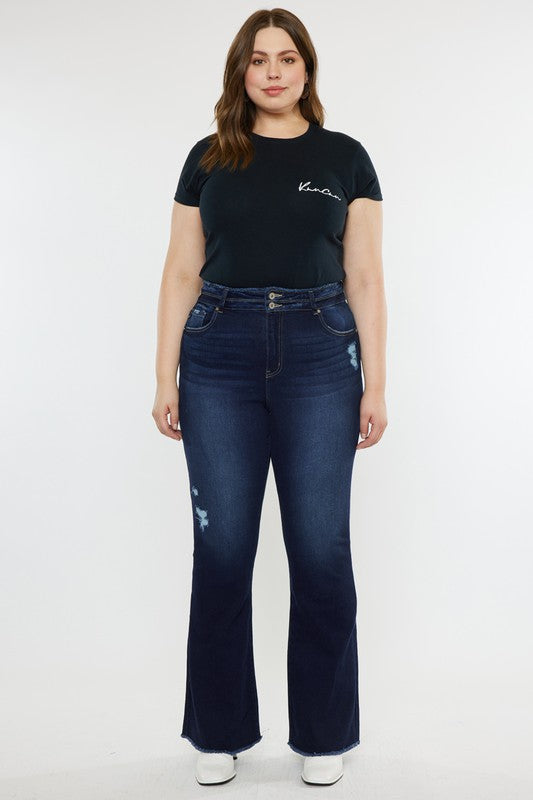 A person stands against a white background, dressed in a black t-shirt and white shoes, showcasing the flattering fit of the Plus High Rise Double WB Fray Hem Flare Jeans in dark blue stretch denim.