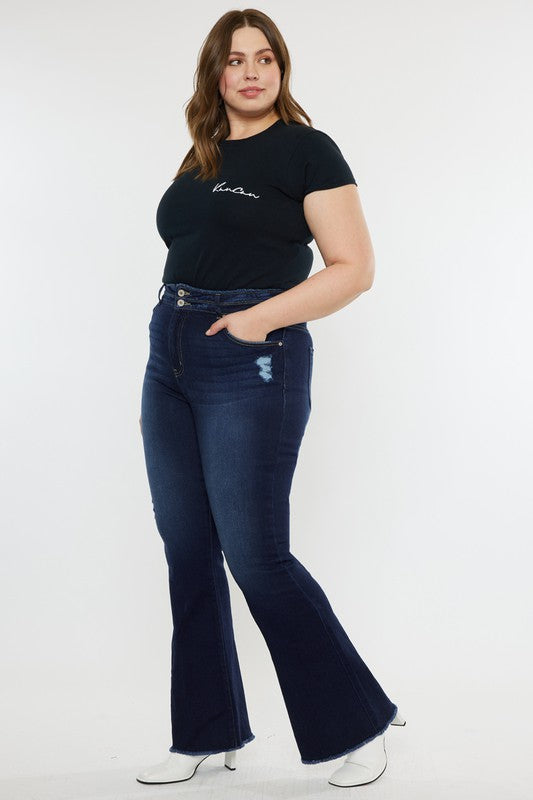 A person stands against a white background, dressed in a black t-shirt and white shoes, showcasing the flattering fit of the Plus High Rise Double WB Fray Hem Flare Jeans in dark blue stretch denim.