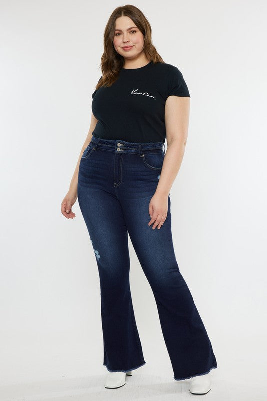 A person stands against a white background, dressed in a black t-shirt and white shoes, showcasing the flattering fit of the Plus High Rise Double WB Fray Hem Flare Jeans in dark blue stretch denim.