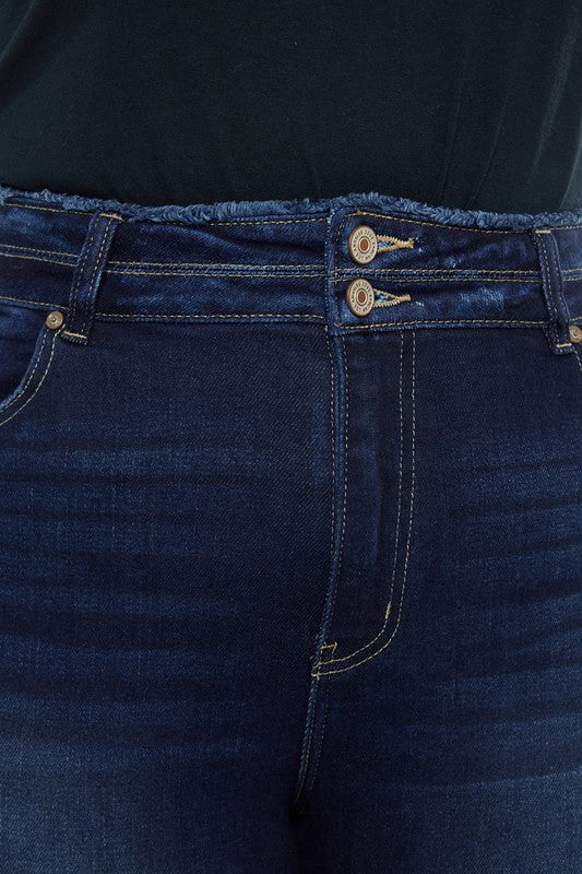 A person stands against a white background, dressed in a black t-shirt and white shoes, showcasing the flattering fit of the Plus High Rise Double WB Fray Hem Flare Jeans in dark blue stretch denim.