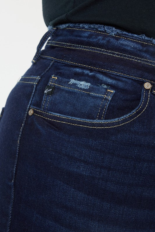 A person stands against a white background, dressed in a black t-shirt and white shoes, showcasing the flattering fit of the Plus High Rise Double WB Fray Hem Flare Jeans in dark blue stretch denim.