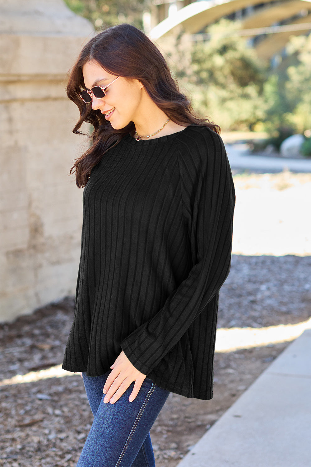 A woman with long brown hair wearing sunglasses and a Basic Bae full-size, slightly stretchy ribbed round neck long sleeve knit top in red, paired with blue jeans, is standing outdoors.