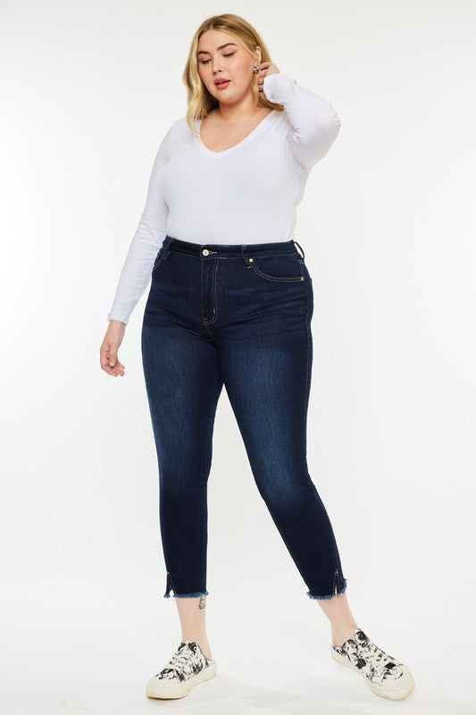 Woman wearing a white long-sleeve shirt, Plus High Rise Ankle Skinny Jeans in dark stone wash, and patterned sneakers stands against a plain white background.