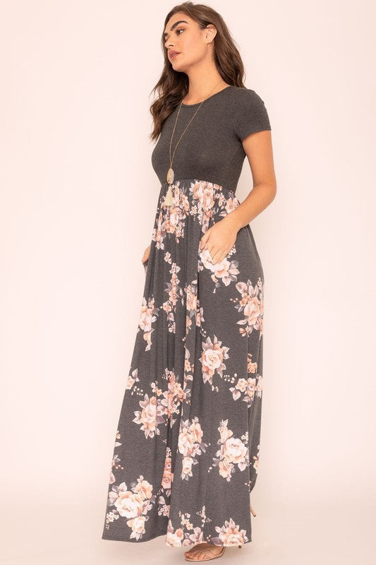 A woman wearing the Plus Short Sleeve Floral Maxi Dress stands smiling, her hands slightly above her waist, against a plain background. Made in the United States, her attire exudes charm and elegance.