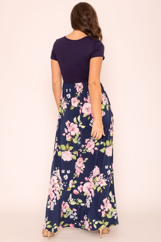 A woman wearing the Short Sleeve Floral Maxi Dress, featuring navy floral designs with pink patterns and a dark top, poses confidently with her hands in the pockets of this US-made outfit.