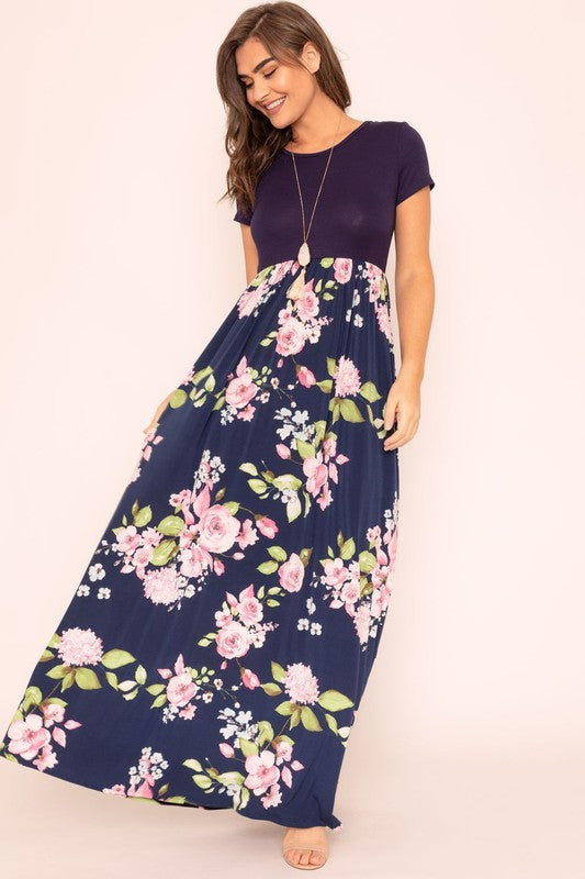 A woman wearing the Short Sleeve Floral Maxi Dress, featuring navy floral designs with pink patterns and a dark top, poses confidently with her hands in the pockets of this US-made outfit.