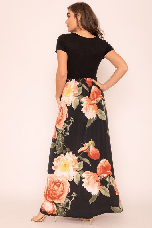 A woman wearing the Short Sleeve Floral Maxi Dress, featuring navy floral designs with pink patterns and a dark top, poses confidently with her hands in the pockets of this US-made outfit.