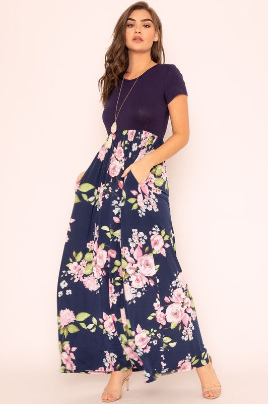 A woman wearing the Short Sleeve Floral Maxi Dress, featuring navy floral designs with pink patterns and a dark top, poses confidently with her hands in the pockets of this US-made outfit.