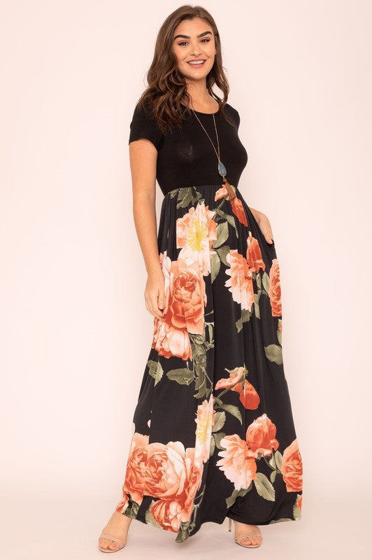 A woman wearing the Short Sleeve Floral Maxi Dress, featuring navy floral designs with pink patterns and a dark top, poses confidently with her hands in the pockets of this US-made outfit.