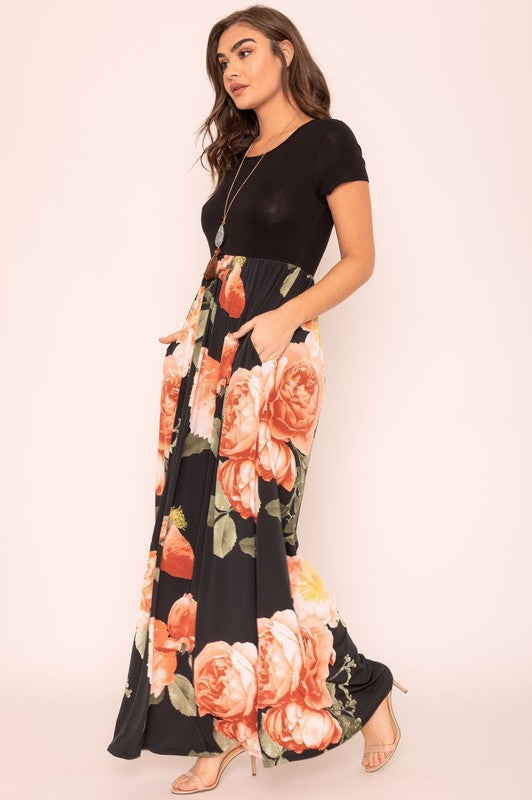 A woman wearing the Short Sleeve Floral Maxi Dress, featuring navy floral designs with pink patterns and a dark top, poses confidently with her hands in the pockets of this US-made outfit.