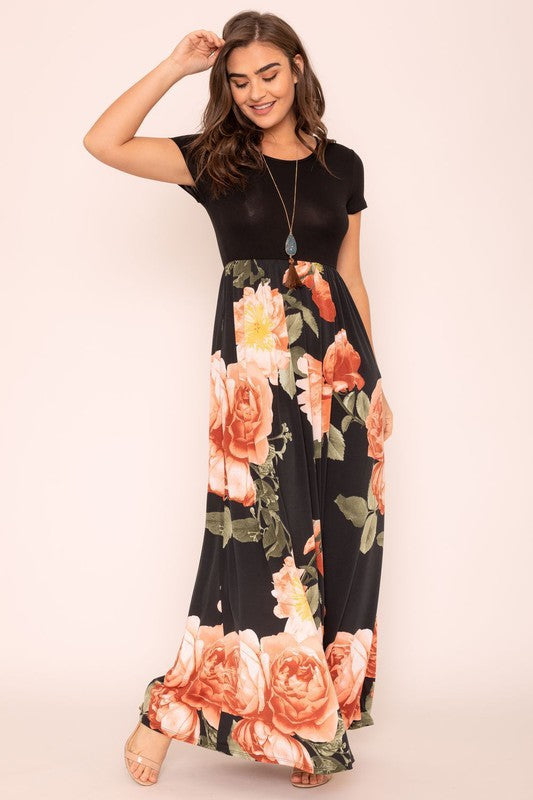 A woman wearing the Short Sleeve Floral Maxi Dress, featuring navy floral designs with pink patterns and a dark top, poses confidently with her hands in the pockets of this US-made outfit.