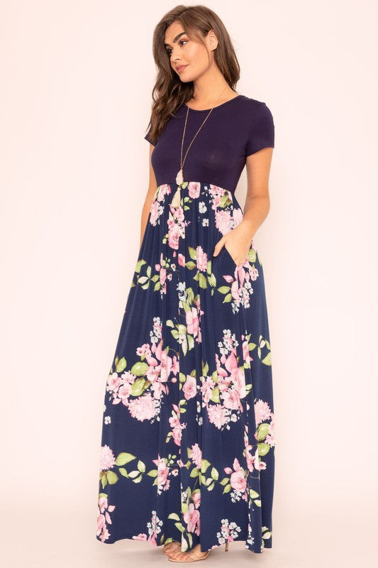 A woman wearing the Short Sleeve Floral Maxi Dress, featuring navy floral designs with pink patterns and a dark top, poses confidently with her hands in the pockets of this US-made outfit.