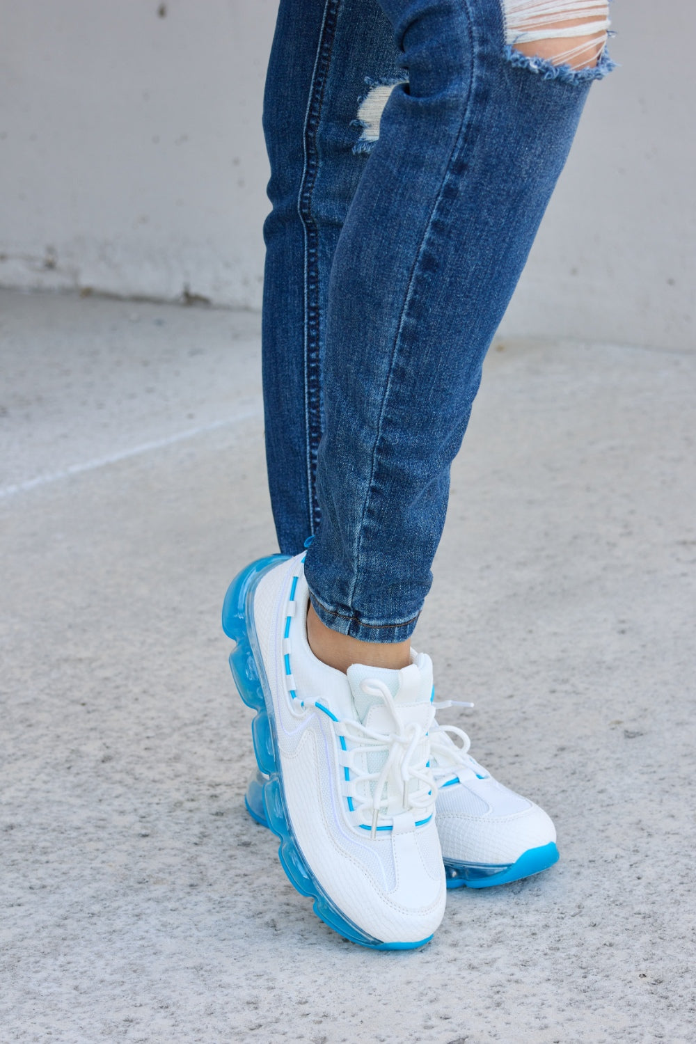 A person wearing ripped jeans and stylish Forever Link Lace-Up Air-Cushioned Athletic Shoes in white with blue accents stands confidently on a concrete surface.