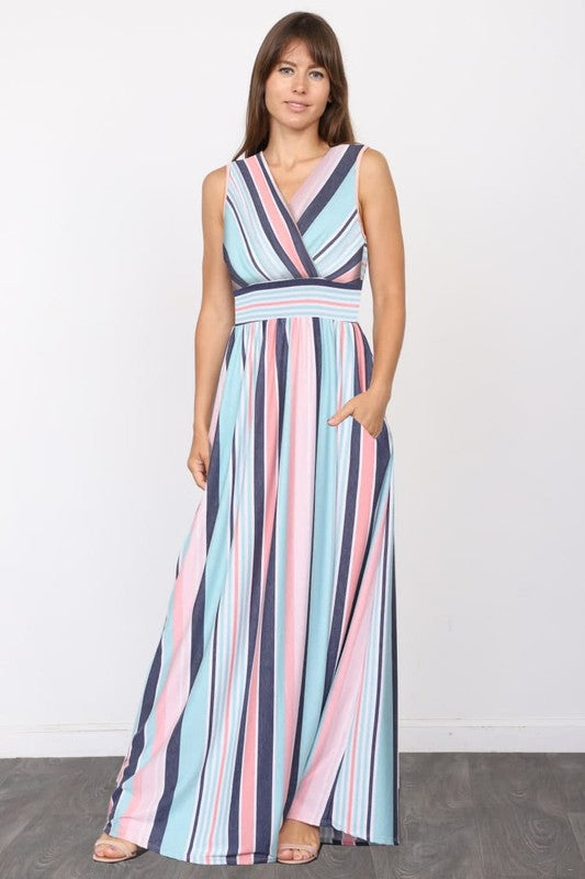 A person wearing the elegant Surplice Stripe Maxi Dress adorned with vertical pastel stripes in blue, pink, and white, stands gracefully on a wooden floor.