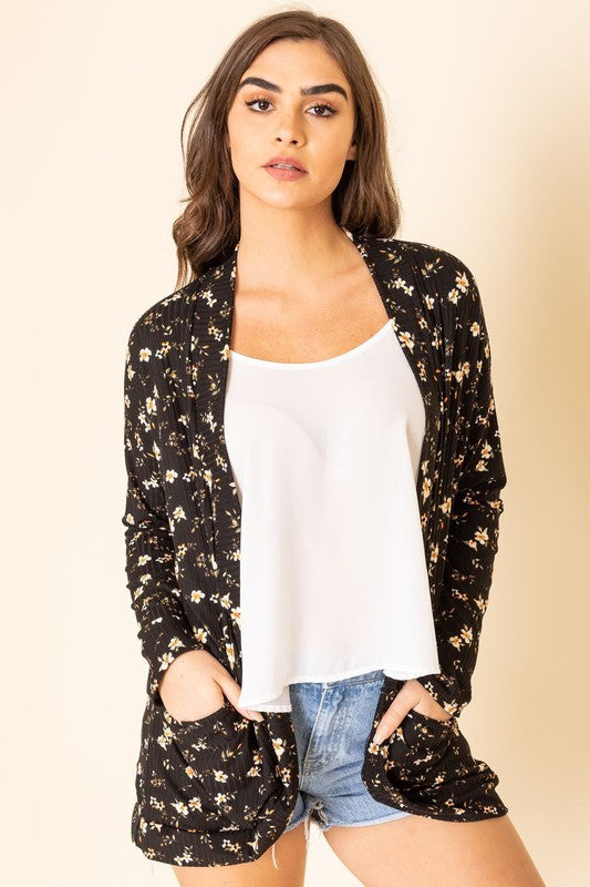 A person wearing a black, polyester-spandex Floral Ribbed Slouch Cardigan pairs it with a white top, denim shorts, and white sneakers while standing against a plain background.