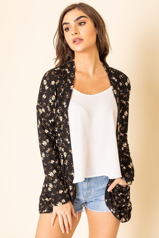 A person wearing a black, polyester-spandex Floral Ribbed Slouch Cardigan pairs it with a white top, denim shorts, and white sneakers while standing against a plain background.