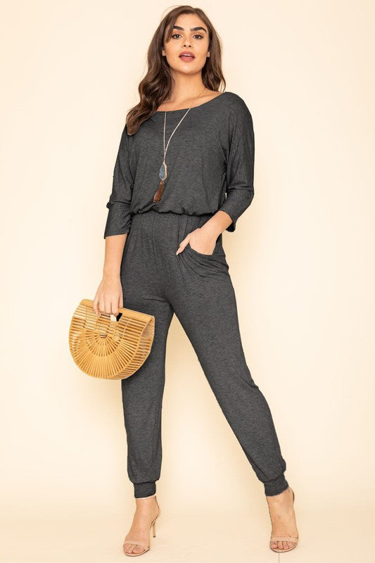 A person in a Quarter Sleeve Boat Neck Blouson Jumpsuit holds a wooden handbag. They are standing against a beige background, wearing a necklace and beige high heels.