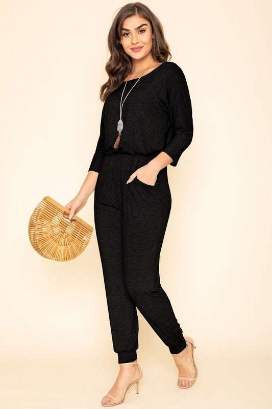 A person in a Quarter Sleeve Boat Neck Blouson Jumpsuit holds a wooden handbag. They are standing against a beige background, wearing a necklace and beige high heels.