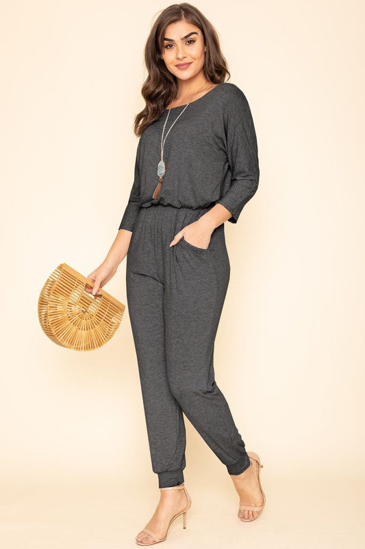 A person in a Quarter Sleeve Boat Neck Blouson Jumpsuit holds a wooden handbag. They are standing against a beige background, wearing a necklace and beige high heels.