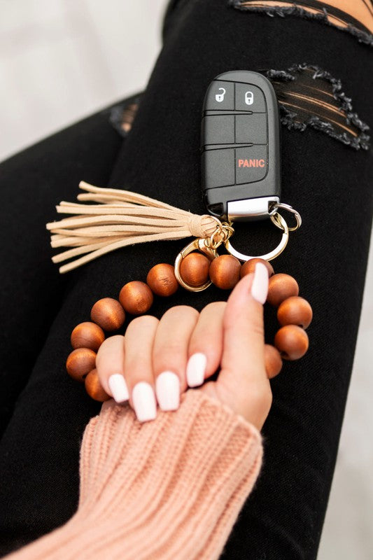 A person holding a car key fob attached to a Wooden Key Ring Bracelet featuring genuine wood beads and a suede tassel rests their hand on their ripped jeans-clad leg.