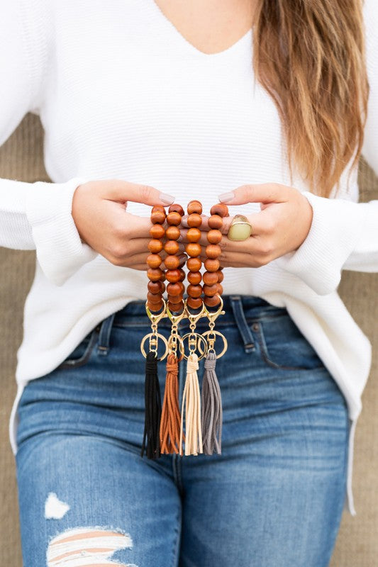 A person holding a car key fob attached to a Wooden Key Ring Bracelet featuring genuine wood beads and a suede tassel rests their hand on their ripped jeans-clad leg.