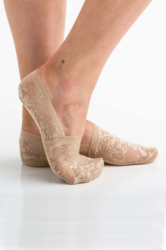 A person wearing No-Slip Floral Lace Socks in beige stands with one foot slightly ahead of the other, revealing two small dot tattoos on the inside of their ankle. The socks showcase a delicate floral lace pattern and have a silicone lining to prevent slipping. The background is plain white.